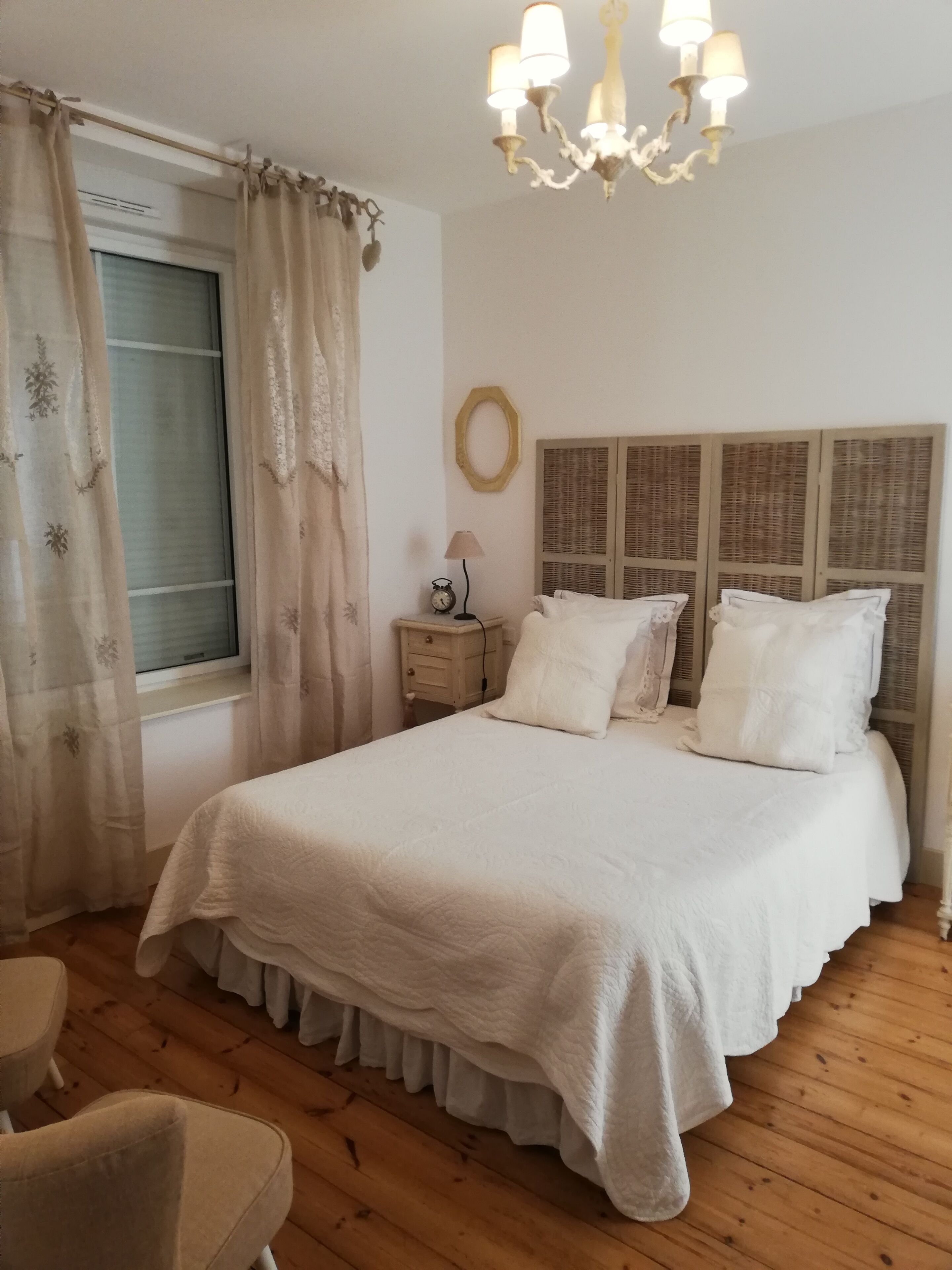 A bedroom with a large bed, a bedside table with a lamp, a window with curtains, and a chandelier.