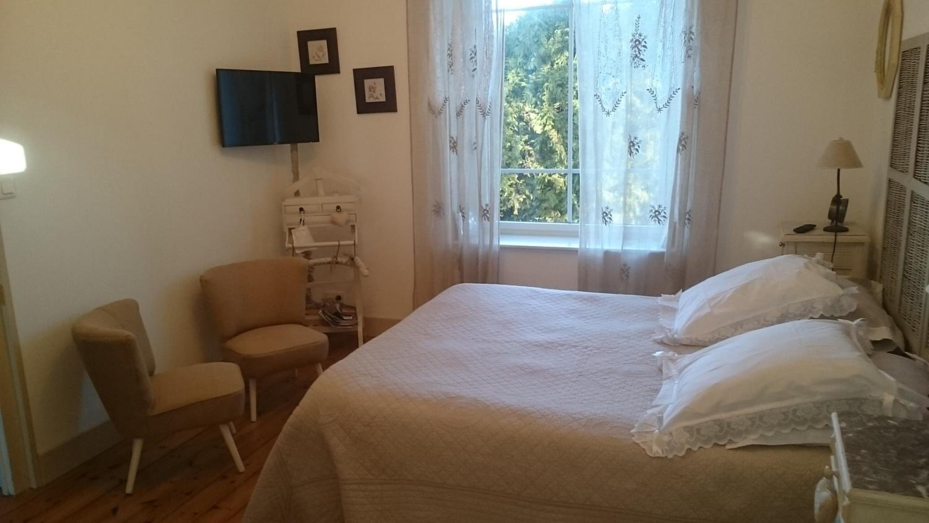 Une chambre à coucher comprenant un lit, une chaise, une table de chevet avec une lampe, un téléviseur fixé au mur et une fenêtre avec des rideaux.