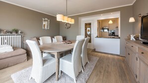 Una cocina moderna con armarios blancos, un comedor con mesa de madera y sillas blancas, y una zona de estar acogedora con un sofá y cojines.