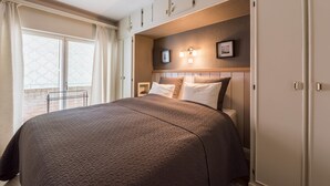 Una cama bien hecha con una colcha texturizada, un cabecero con luces fijadas a la pared y una ventana con cortinas.