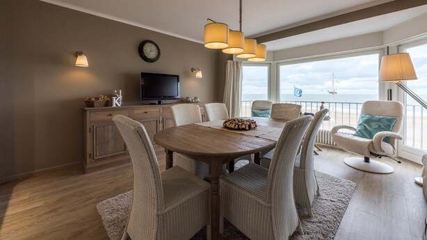 Un comedor con mesa de madera y sillas de mimbre, televisor y vista al océano.