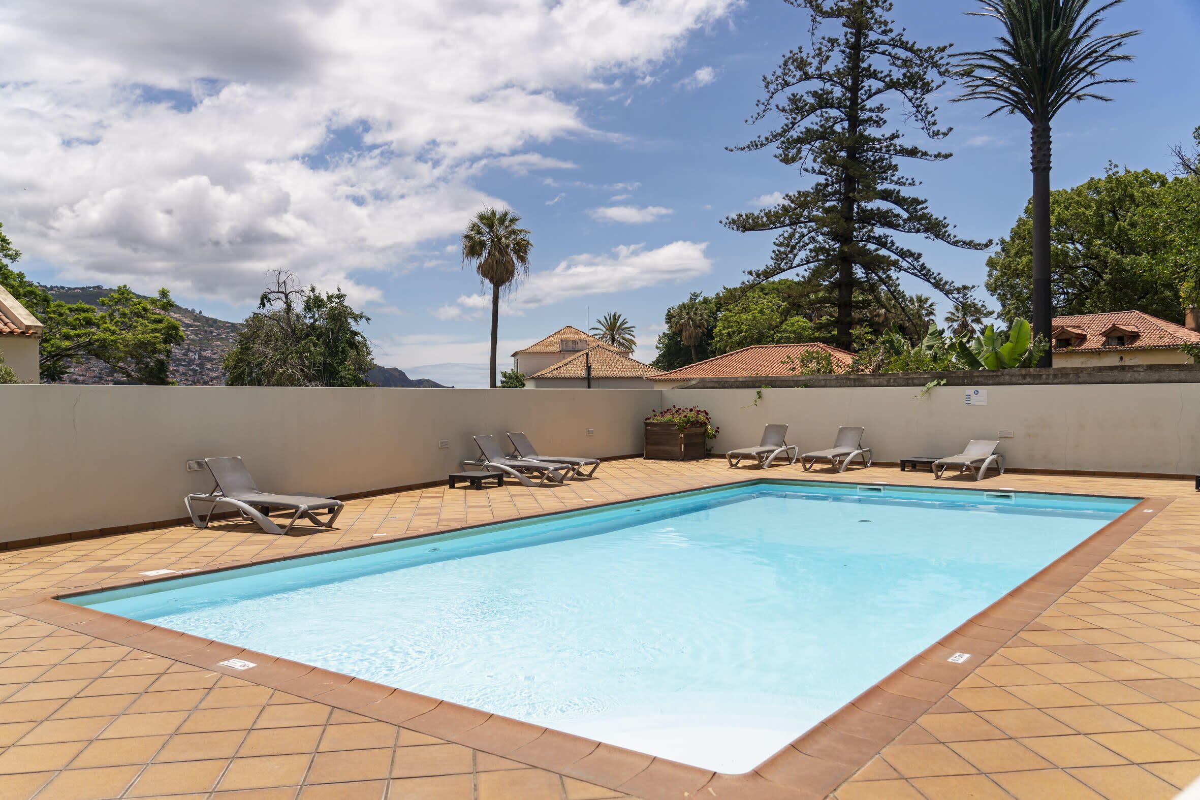 A rectangular swimming pool with tiled edges, surrounded by lounge chairs and a tiled deck.