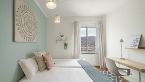 A modern bedroom with two beds, a desk, a view of the city, and a decorative wall piece.