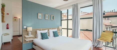 A modern hotel room with a large bed, a view of the city, and a balcony with a chair.