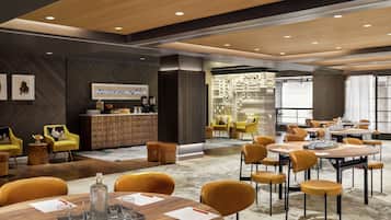 A modern hotel lobby with round tables, yellow chairs, and a bar area.
