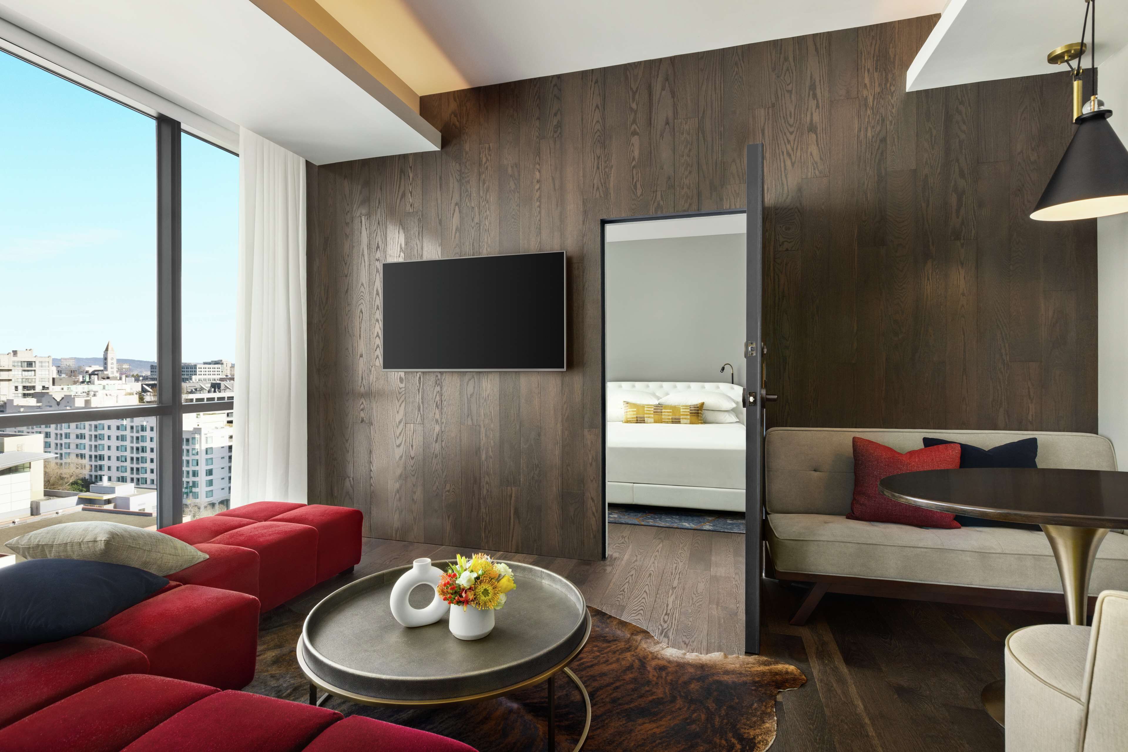 Een moderne hotelkamer met een groot raam, een flatscreen televisie, een bank, een rode fauteuil, een klein rond tafeltje met een bloevas, en een houten vloer.