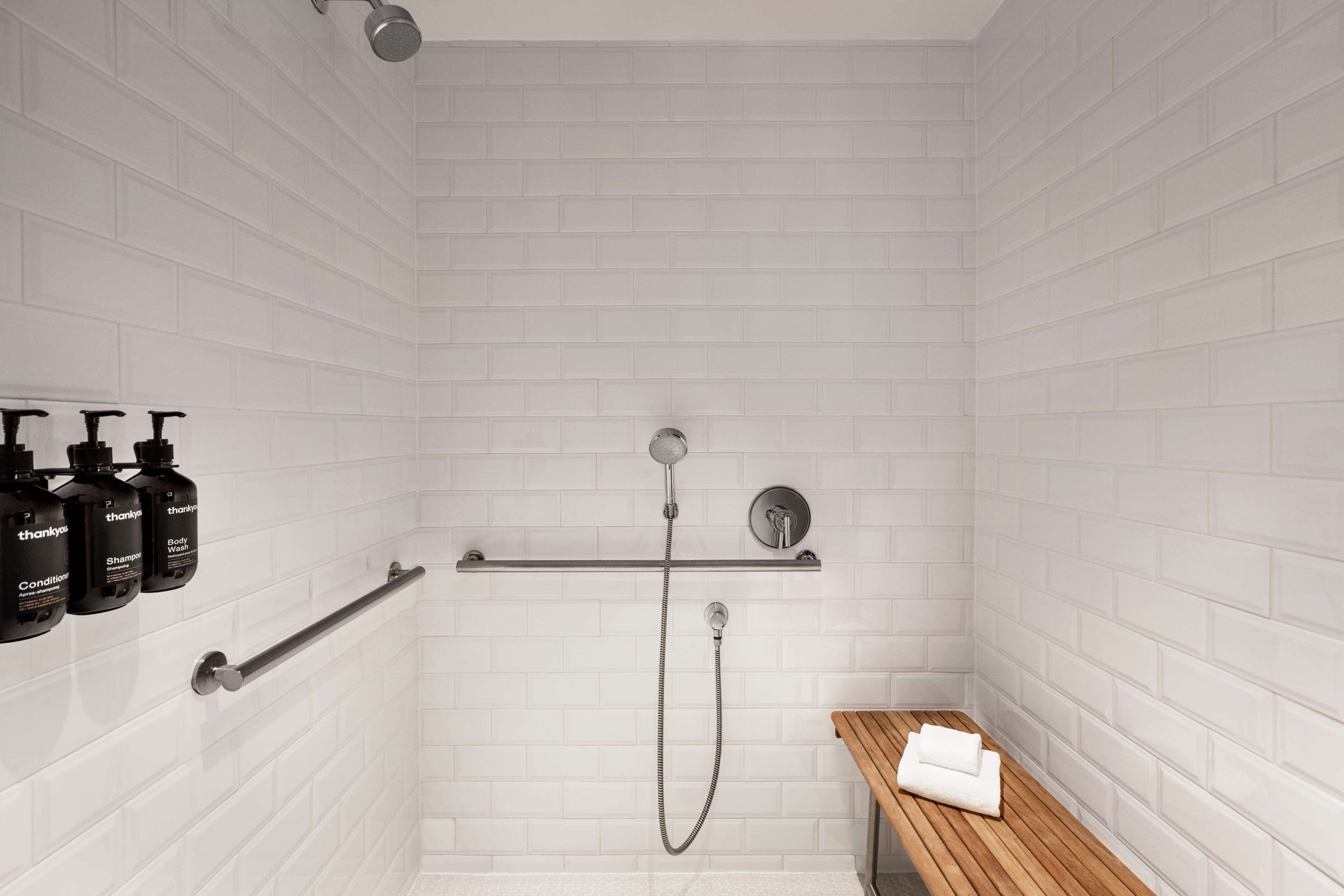 A modern shower area with white tiled walls, a wooden bench, and a shelf with toiletries.