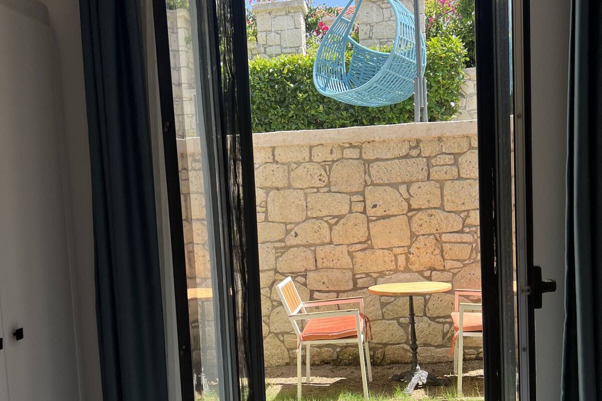 A room with a bed, a sliding glass door leading to an outdoor area with a stone wall, a blue hanging chair, and two chairs with a table.