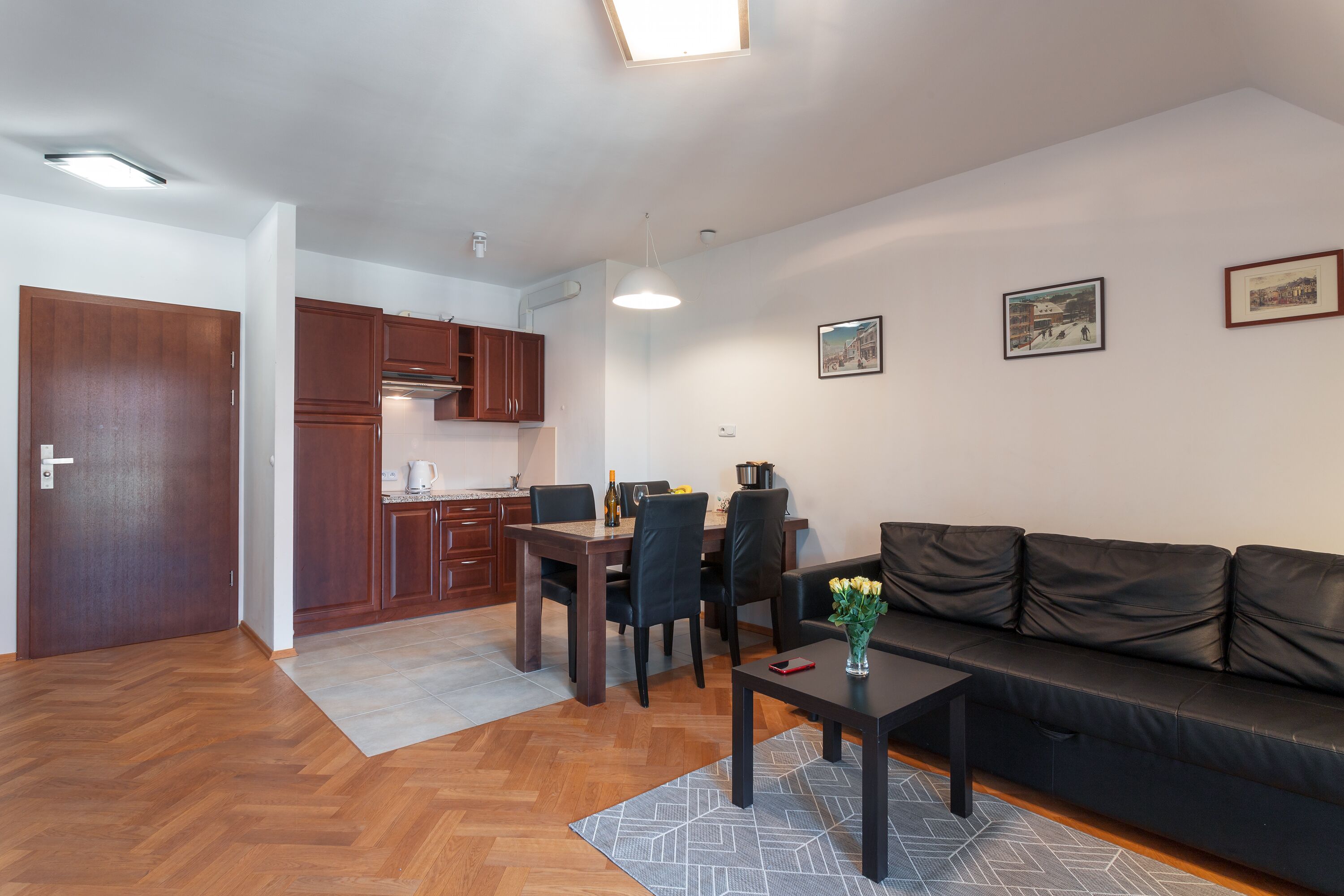 A modern living room with a black leather sofa, a dining table with chairs, a kitchen area with wooden cabinets, and a hallway.