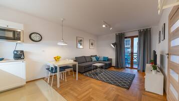 A modern living room with a sofa, coffee table, and dining area. There is a kitchen with a microwave and a clock on the wall.