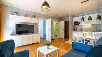 Una sala moderna con un televisor de pantalla plana, un sofá, una mesa de centro con una planta y una zona de comedor con una mesa y sillas blancas.