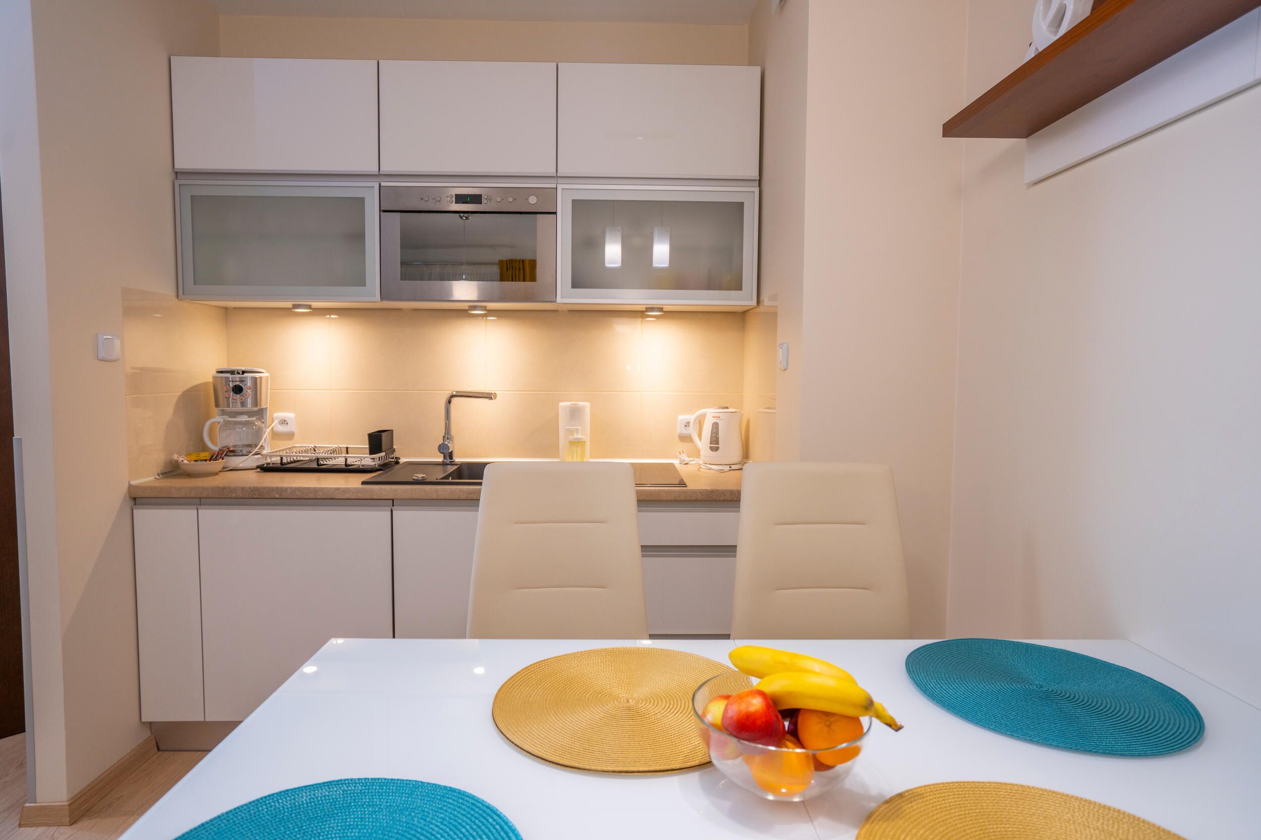 A modern kitchen with white cabinets, a microwave, and a coffee maker. There is a dining table with a placemat and a bowl of fruit.