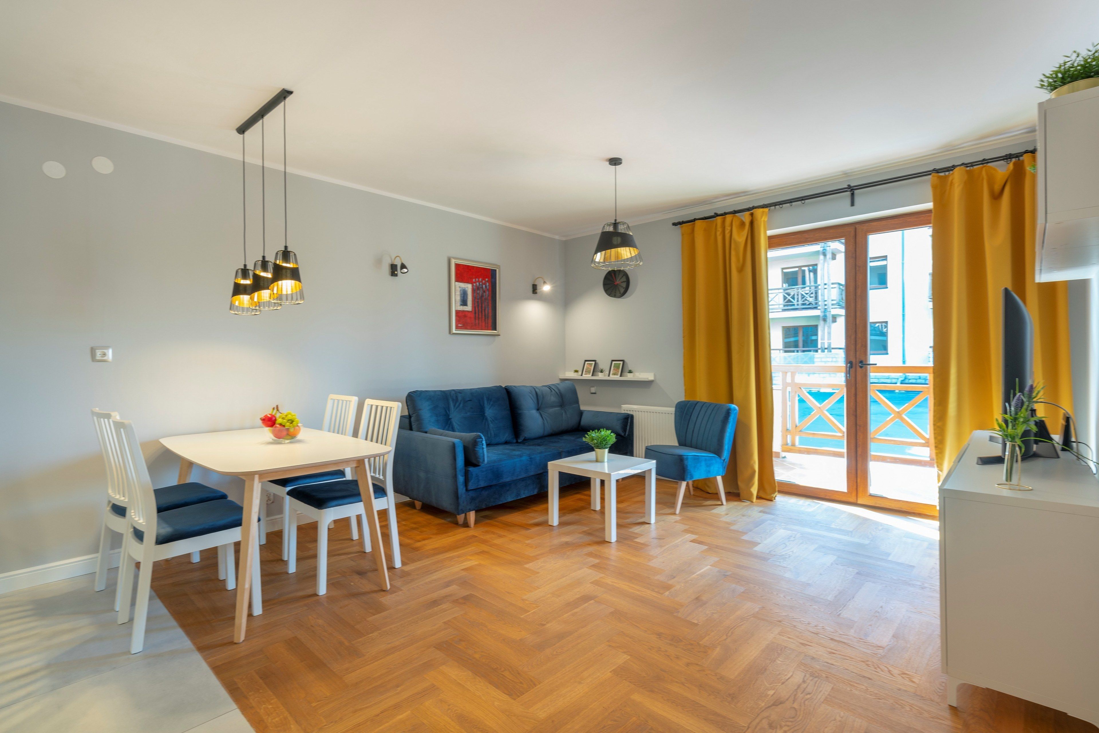 A modern living room with a blue sofa, white dining table, and chairs, a small white side table, and a hanging light fixture.