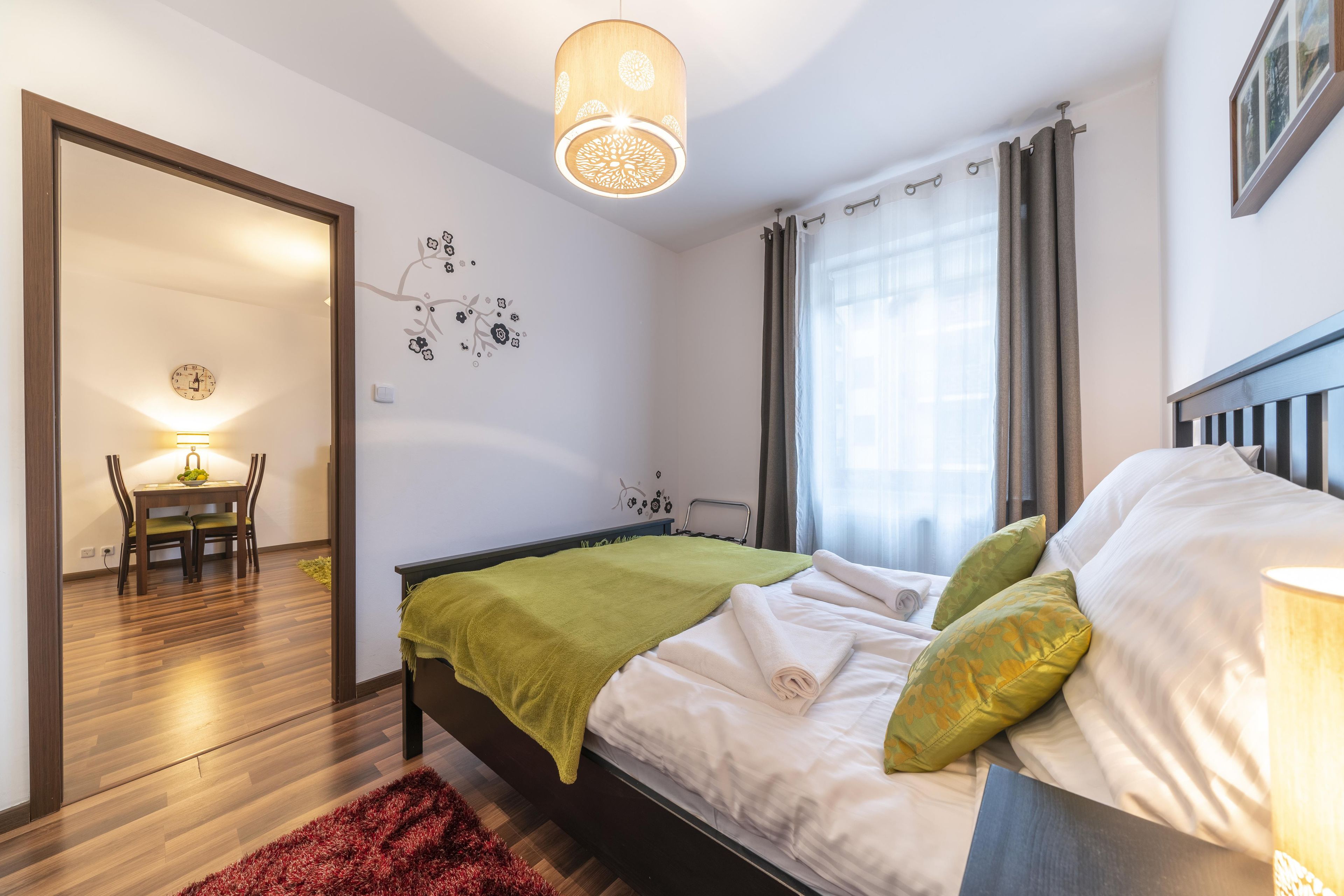 A bedroom with a bed, green pillows, a wooden headboard, a red rug, a wall clock, and a dining area with a table and chairs.