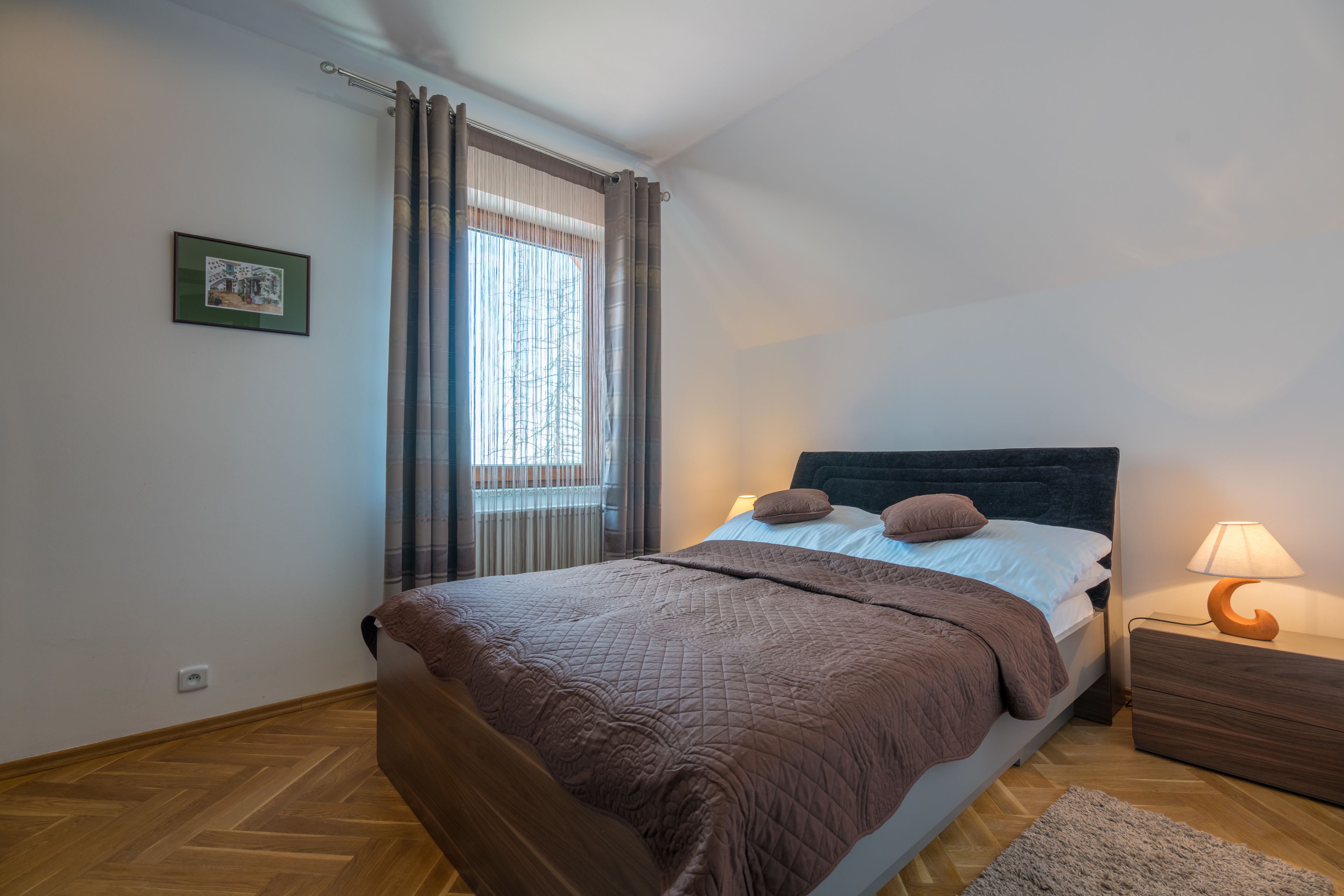 A bedroom with a bed, a nightstand, a lamp, a window with curtains, and a framed picture on the wall.