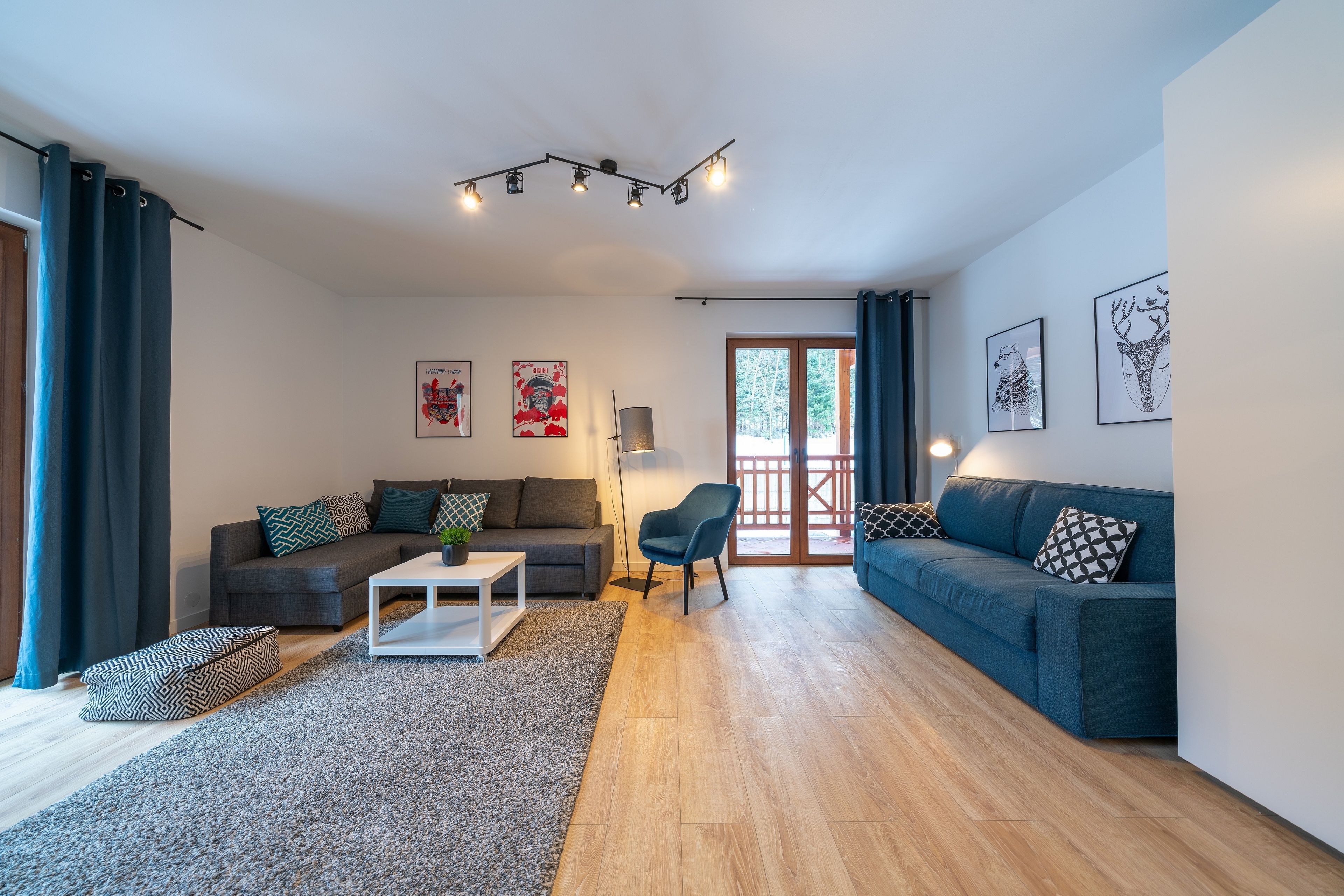 Una sala de estar moderna con un sofá gris, un sofá azul, una mesa de centro blanca y un pouf estampado. Hay dos cuadros enmarcados en la pared, una lámpara de pie y un ventanal con cortinas.