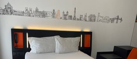 A hotel room with a large bed, white pillows, an orange accent, a cityscape wall art, a bedside phone, and a desk with a chair.