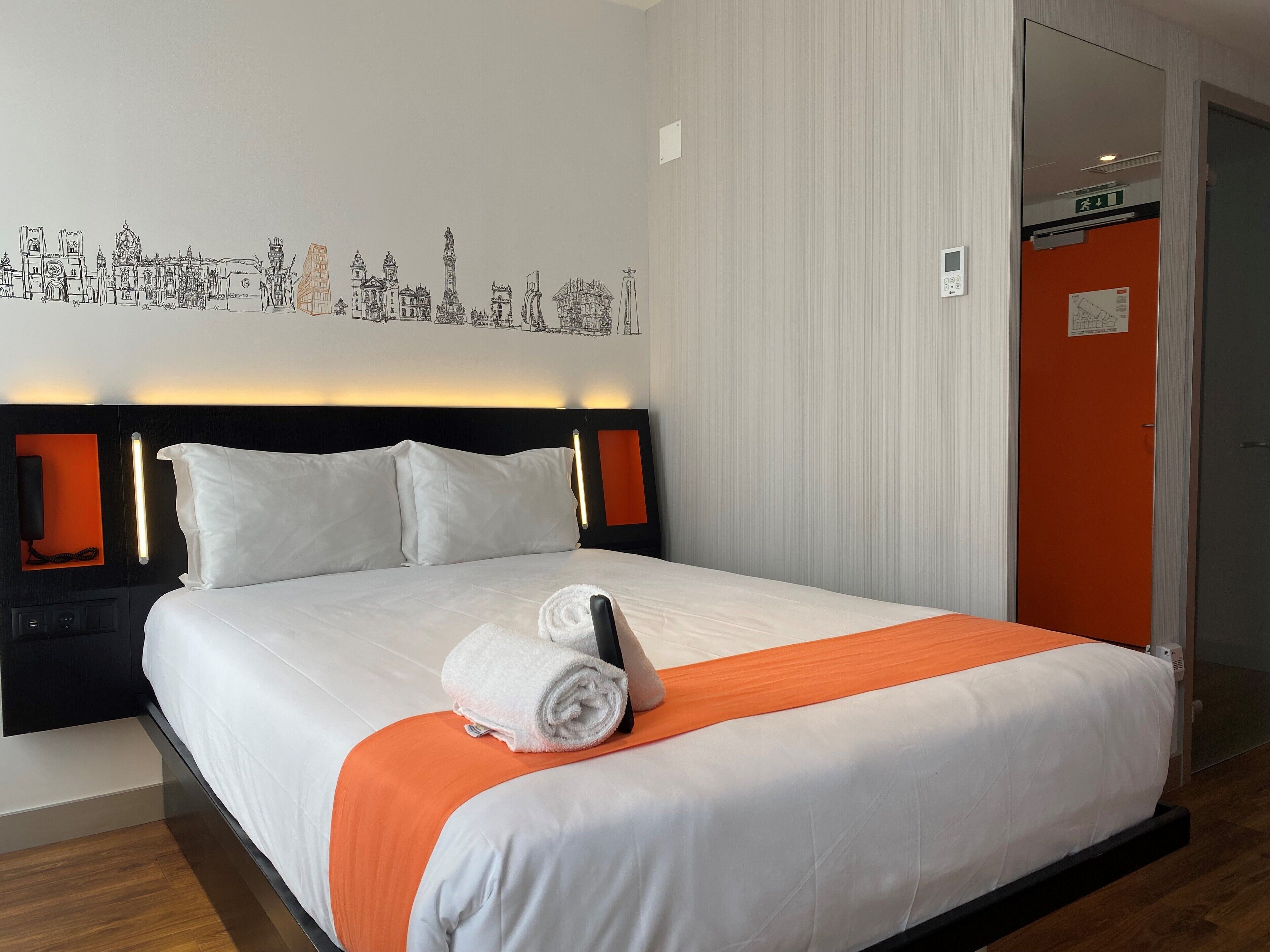 Una habitación de hotel con una cama bien hecha, un mural de un paisaje urbano en la pared y una puerta con detalle naranja.