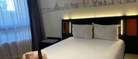 A hotel room with a large bed, a headboard with a cityscape mural, a bedside table with a lamp, and a window with sheer curtains.