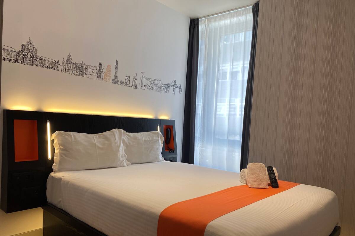 Une chambre d’hôtel avec un grand lit, une tête de lit dotée d’une bande lumineuse, une table de chevet avec un téléphone et une fenêtre ornée de rideaux légers.