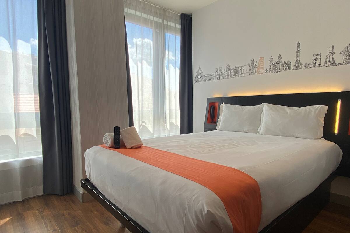 Une chambre d’hôtel avec un grand lit, une décoration murale représentant un paysage urbain et une vue sur l’extérieur par la fenêtre.