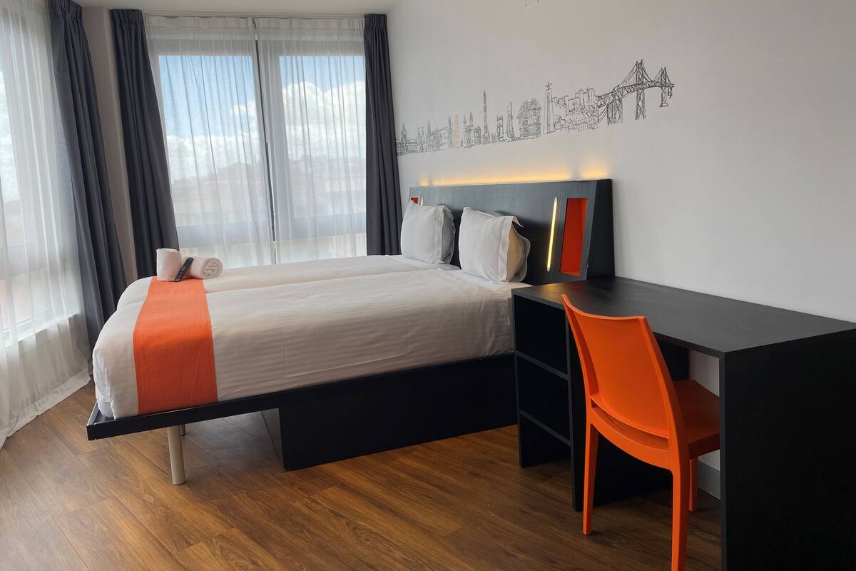 Une chambre d’hôtel avec un lit, un bureau et une chaise. Il y a une fenêtre avec des rideaux et un mur orné d’une fresque représentant un paysage urbain.