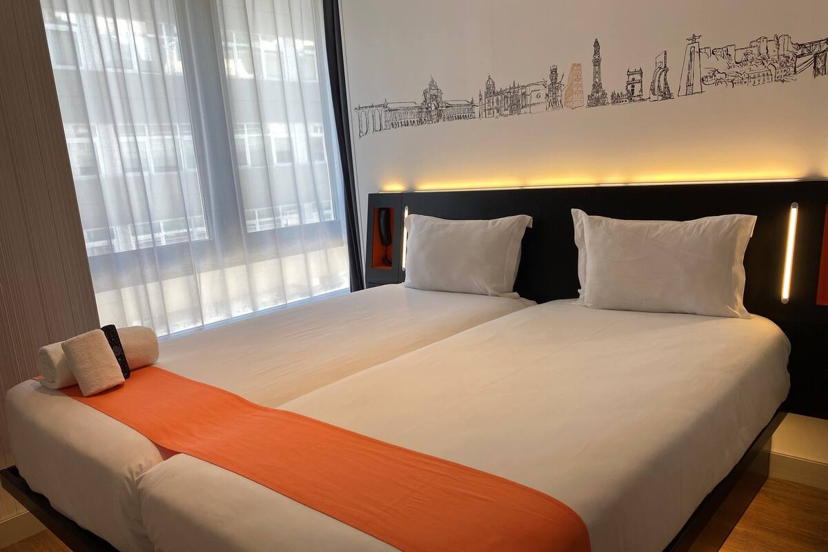Un lit double avec du linge de lit blanc et une touche de couleur orange, une table de chevet avec un téléphone, et une fresque représentant un paysage urbain sur le mur.