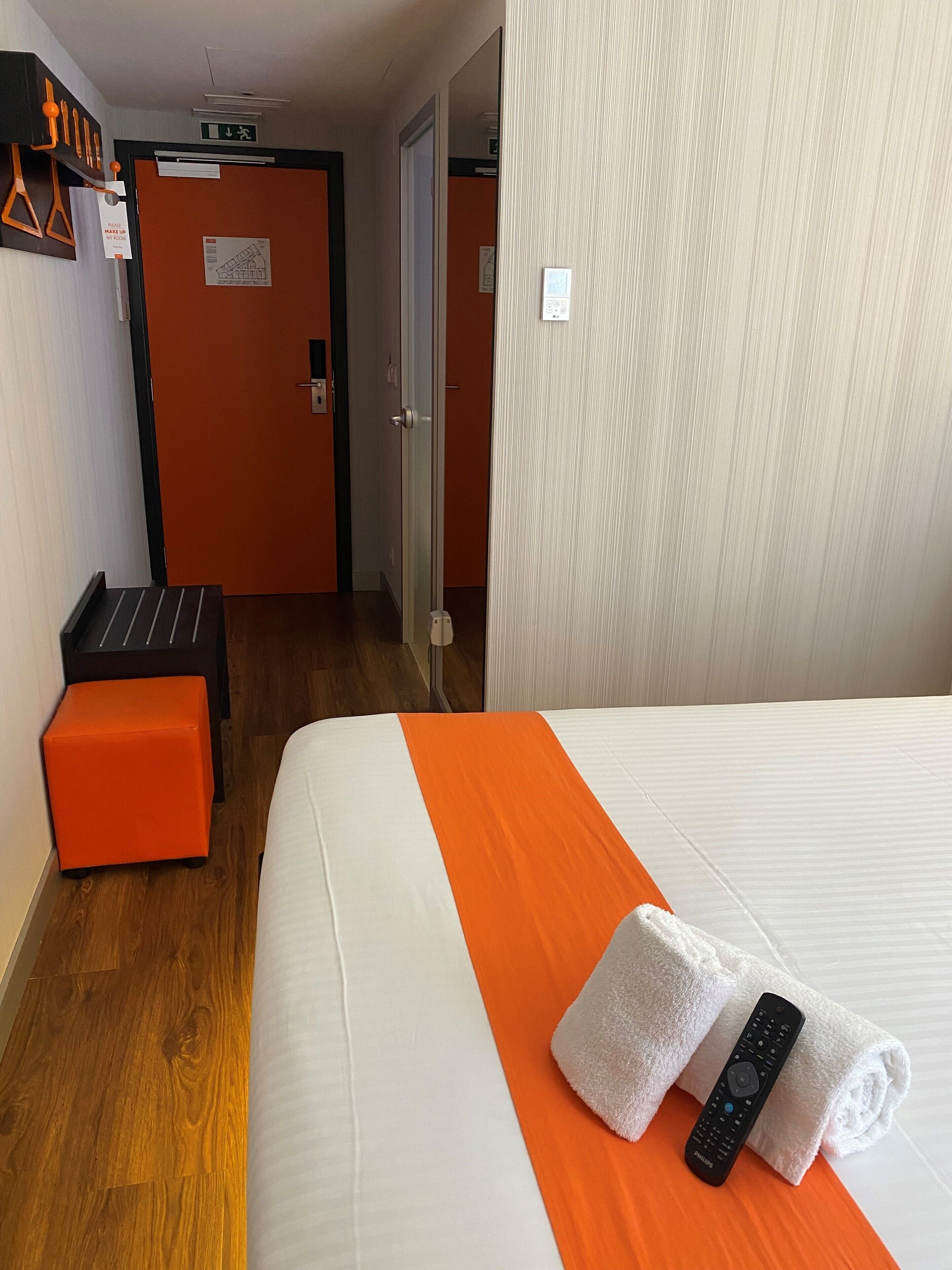 Una habitación de hotel con una cama, un pequeño taburete naranja, un control remoto y una toalla enrollada.