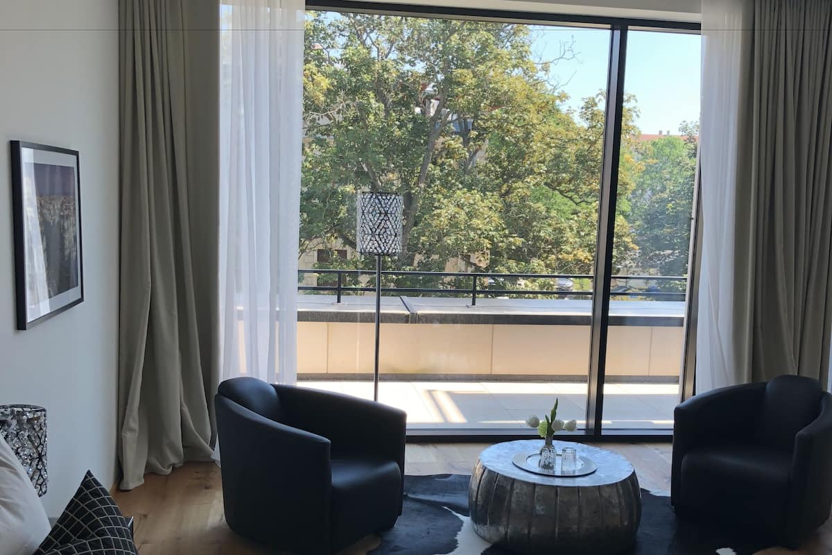 Quarto de hotel moderno com uma cama grande, duas poltronas pretas, uma mesa de centro redonda e vista para árvores e quadra de basquete através de janelas amplas.