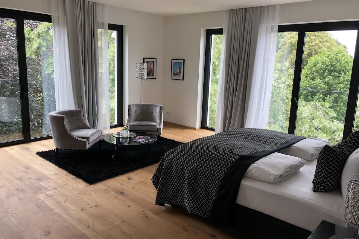 Quarto moderno com uma cama grande, duas cadeiras, uma mesa de centro e janelas amplas com vista para o verde.