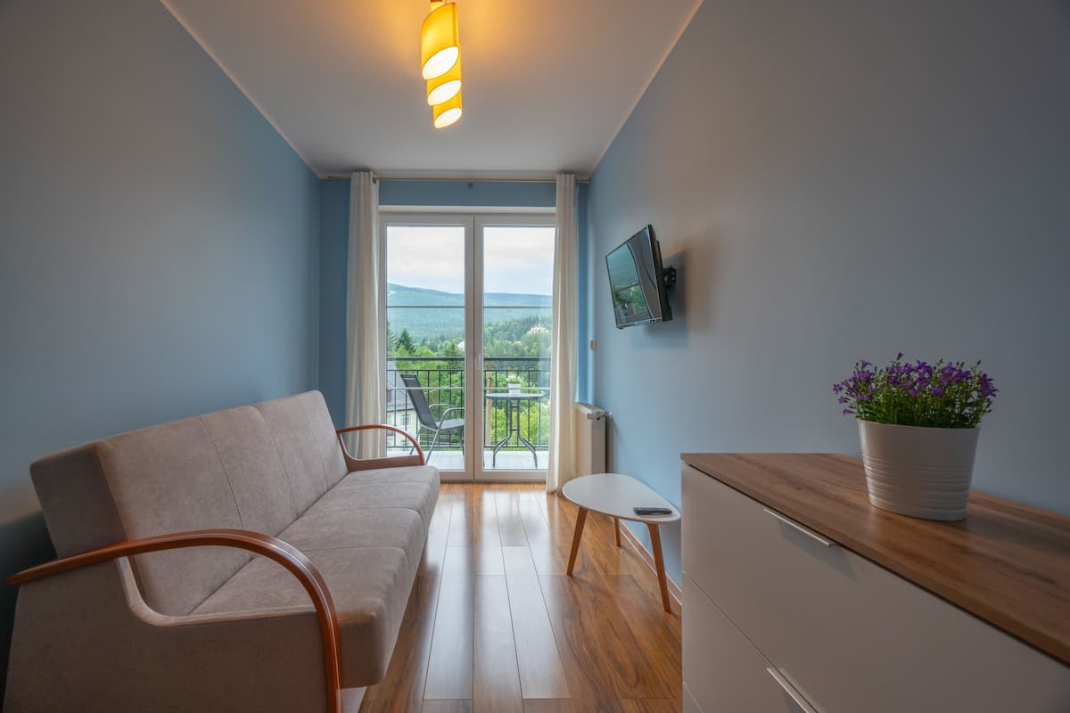 Una sala de estar moderna con sofá, una mesita, televisor montado en la pared y un balcón con vistas a zonas verdes.