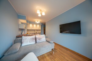 A modern living room with a grey sofa, a flat-screen TV mounted on the wall, and a kitchen area in the background.