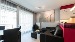 A modern living room with a black sofa, a dining table with chairs, and a large window with blinds. - Jersey 201 (Koksijde)