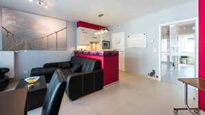 A modern living room with a black leather sofa, a dining table, and a kitchen area with a red accent wall. - Jersey 201 (Koksijde)