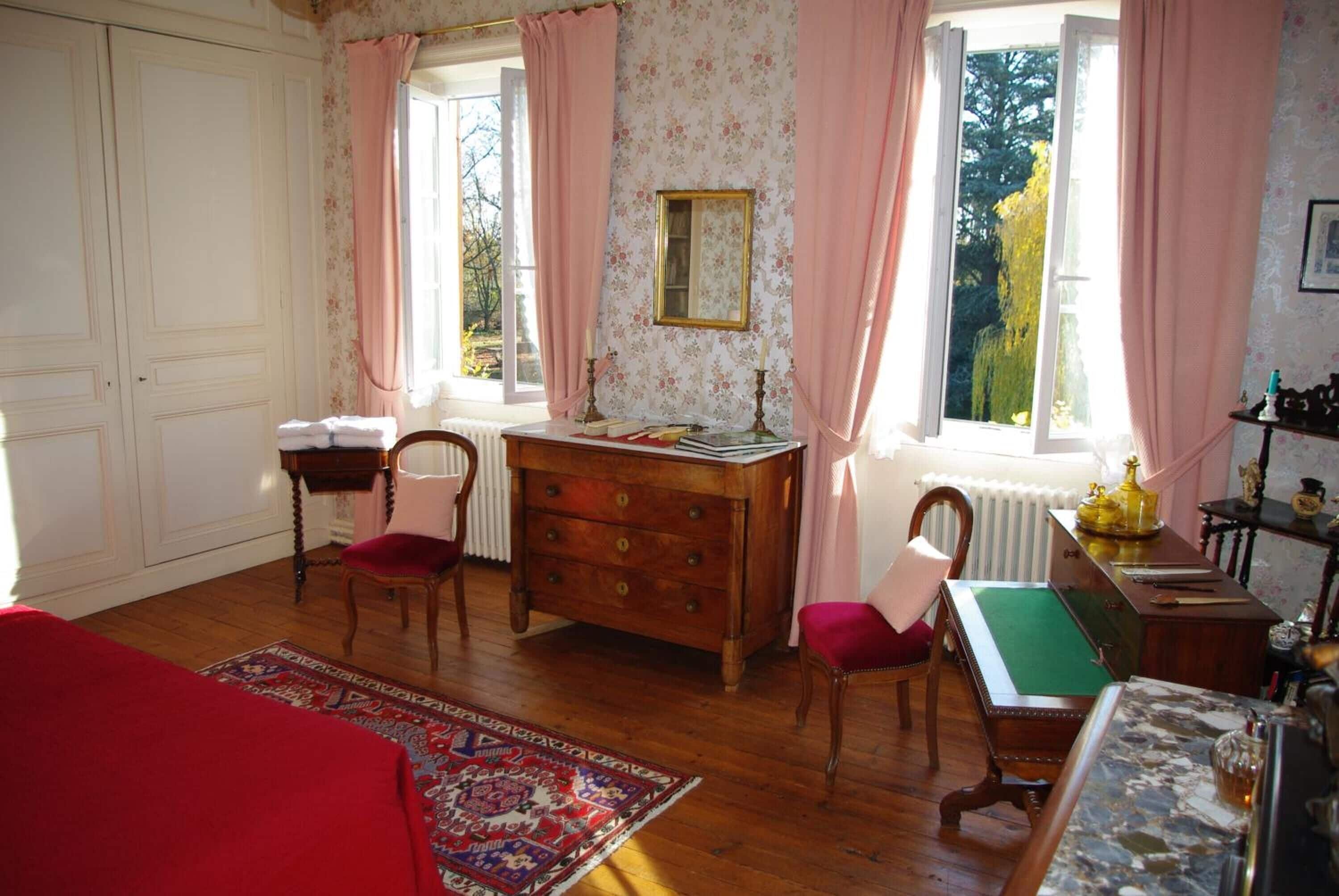 Una habitación con piso de madera, un sofá rojo, un escritorio con cajones, una silla, una ventana con cortinas rosadas y vista a árboles desde el exterior.