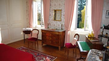 Una habitación con piso de madera, un sofá rojo, un escritorio con cajones, una silla, una ventana con cortinas rosadas y vista a árboles desde el exterior.