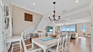 A spacious dining area with a white table and chairs, a chandelier, and a staircase leading to another level. - DIRECT GULF FRONT LUXURY HOME, INCREDIBLE BEACHES, SPACIOUS LAYOUT. (Gulf Shores)