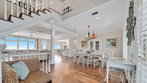 A spacious living area with a wooden floor, a white dining table with chairs, a wicker sofa with cushions, and a staircase leading to an upper level. - DIRECT GULF FRONT LUXURY HOME, INCREDIBLE BEACHES, SPACIOUS LAYOUT. (Gulf Shores)