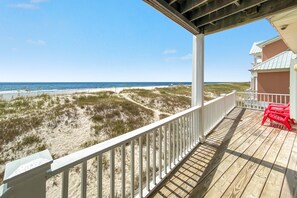 Eine Holzterrasse mit weißer Reling, die auf einen Strand mit Dünen und dem Meer blickt.