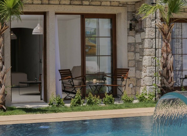 A poolside area with a stone wall, two wooden chairs, a glass table, and a hanging light fixture.