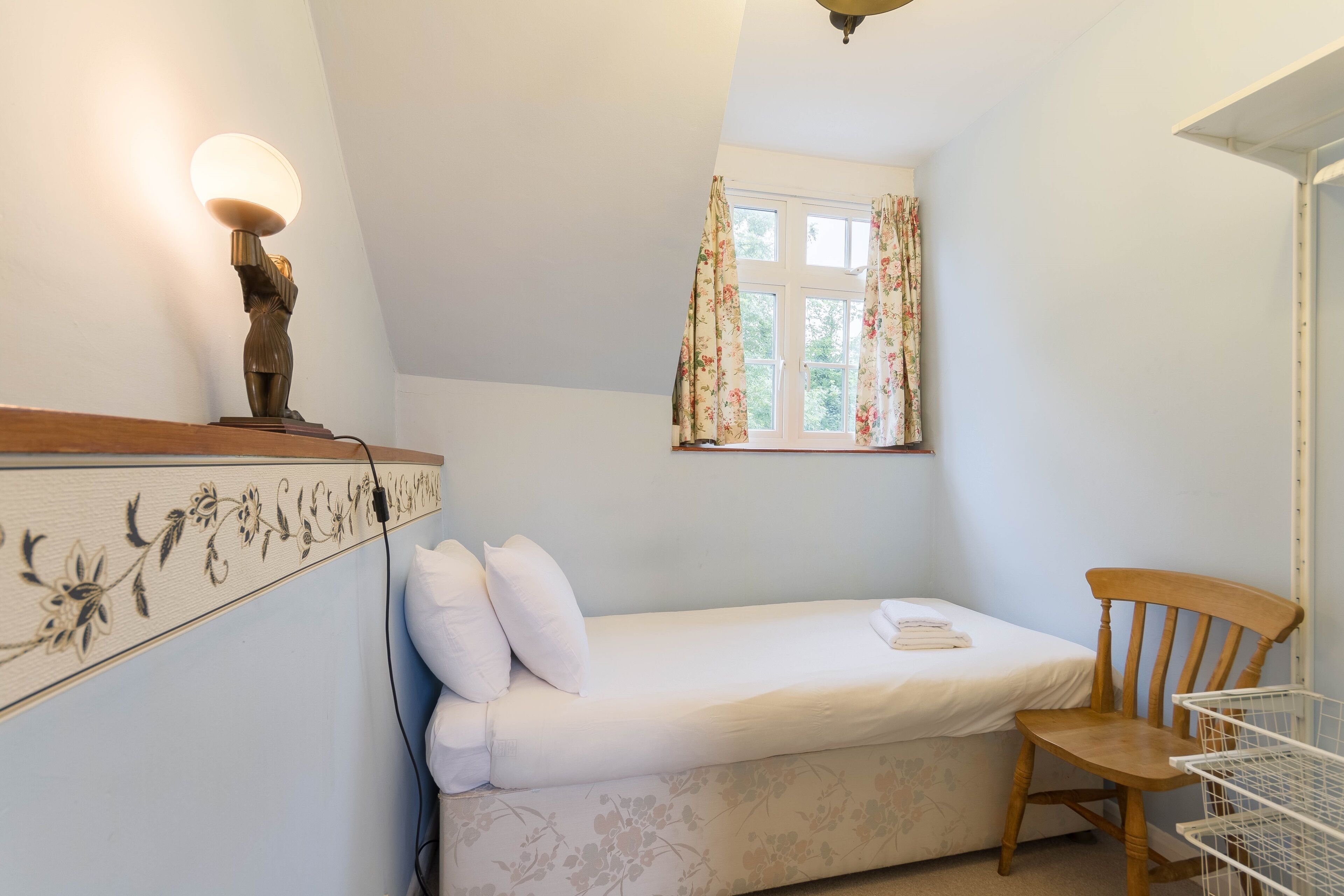 A small, neatly arranged bedroom with a single bed, a wooden chair, a bedside table with a lamp, and a window with floral curtains.