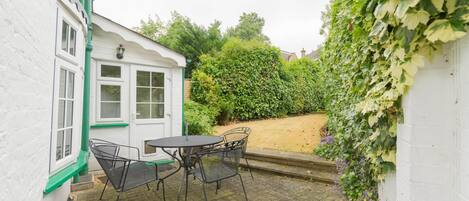 A patio with a table and chairs, a white building with green trim, and a hedge.