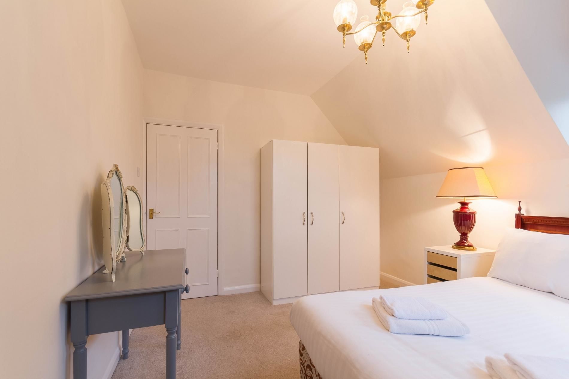 A bedroom with a bed, a nightstand with a lamp, a wardrobe, and a mirror.