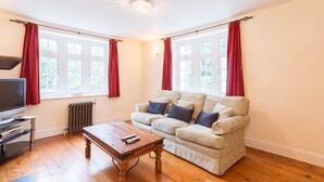 Una sala de estar con sofá, mesa de centro, televisor y ventanas grandes con cortinas rojas.