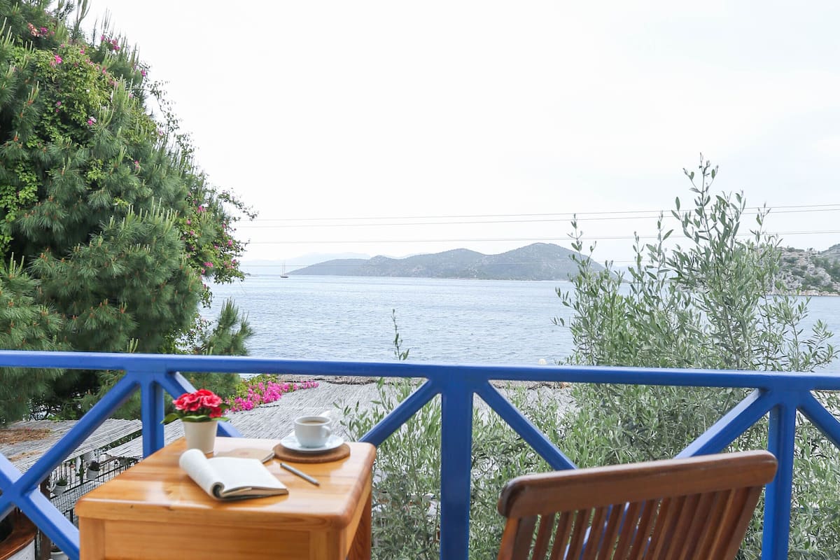 Een houten tafel met een stoel en een boek, een kopje en een bloem op een balkon met een blauwe reling, uitkijkend op een wateroppervlak en heuvels.