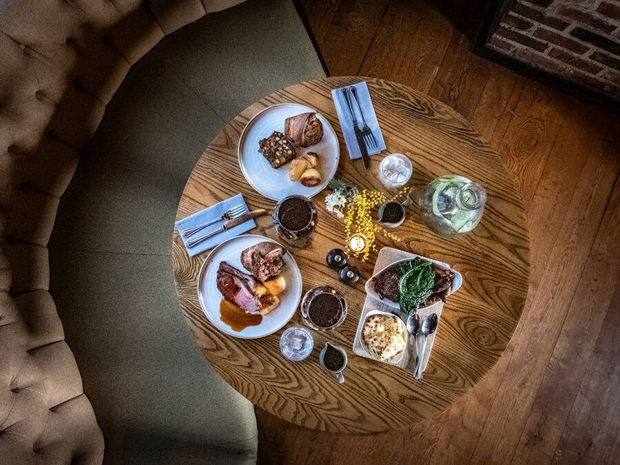 Una mesa redonda de madera con diversos platos y condimentos, rodeada por un sofá capitonado.