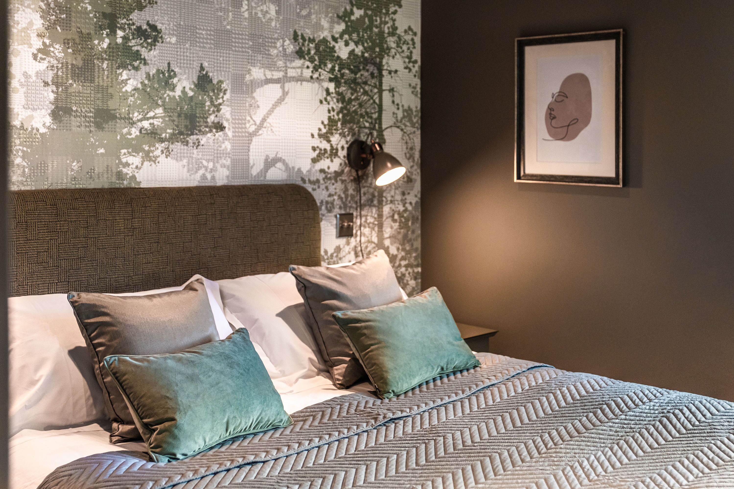 A neatly made bed with patterned bedding, a wall-mounted lamp, and a framed artwork.