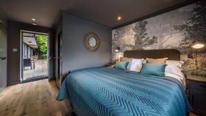 A bedroom with a large bed, a wall-mounted mirror, and a view of the outdoors through an open door. - The Cricketers (Saffron Walden)