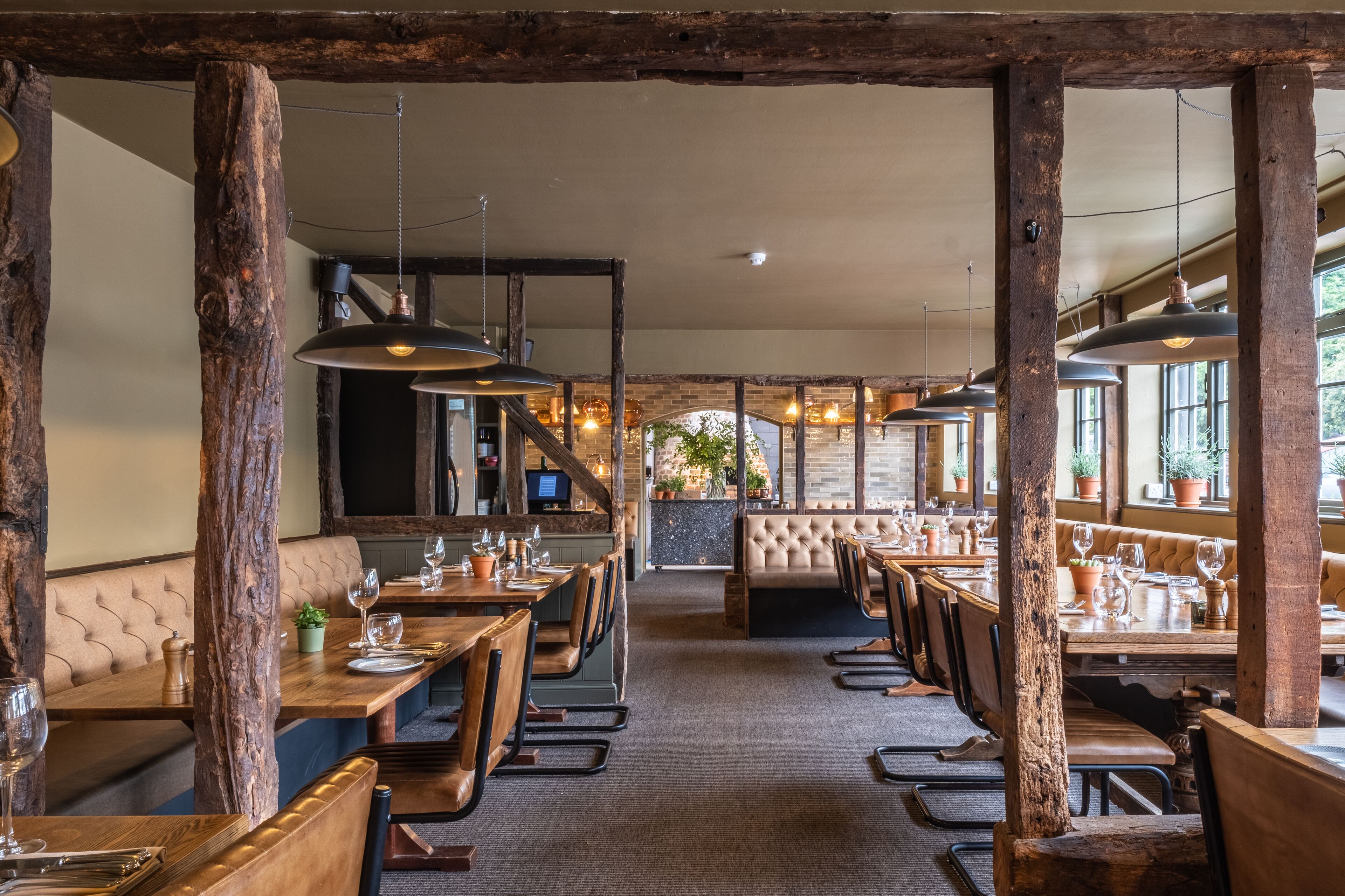 A restaurant with wooden beams, leather booths, and hanging pendant lights. Tables are set with wine glasses and cutlery.