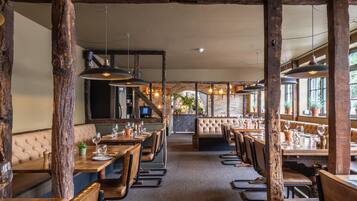 A restaurant with wooden beams, leather booths, and hanging pendant lights. Tables are set with wine glasses and cutlery.